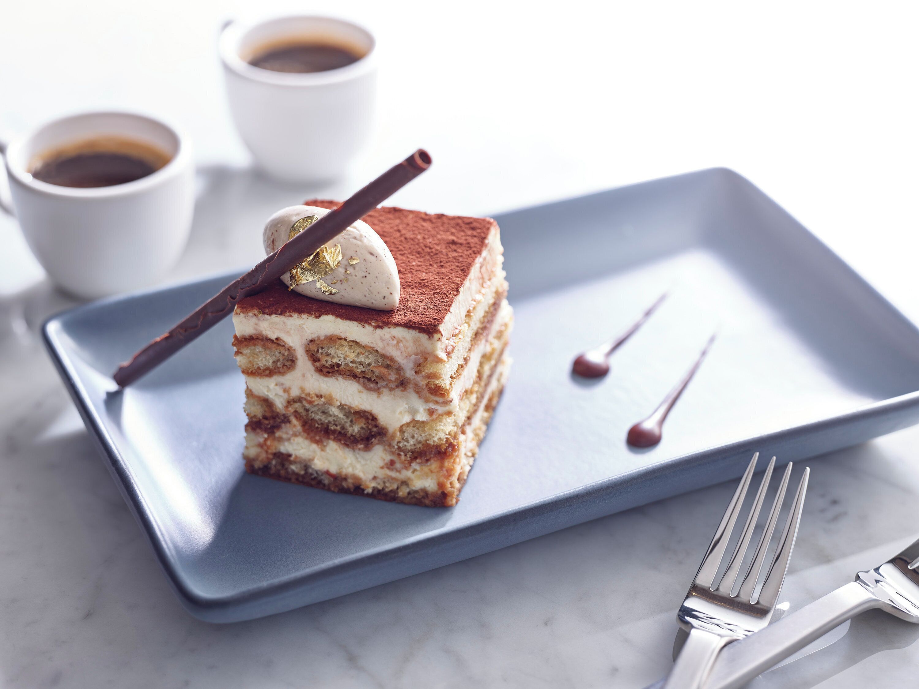 Eine Scheibe mehrschichtiger Dessert auf blauem Teller, mit einer Gabel und zwei Tassen Kaffee im Hintergrund.