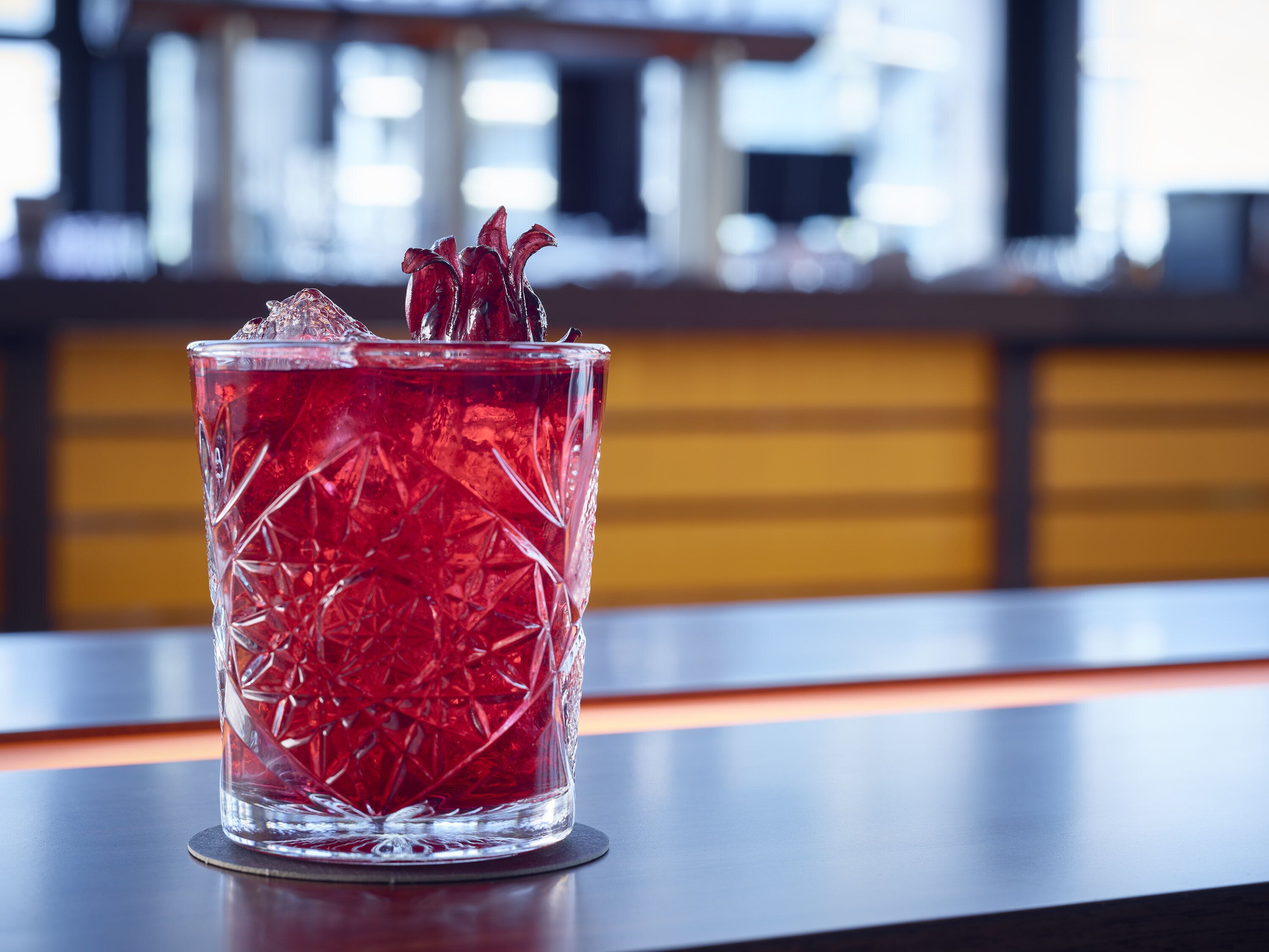 Un vaso transparente con una bebida roja, cubitos de hielo y una flor decorativa sobre una barra.