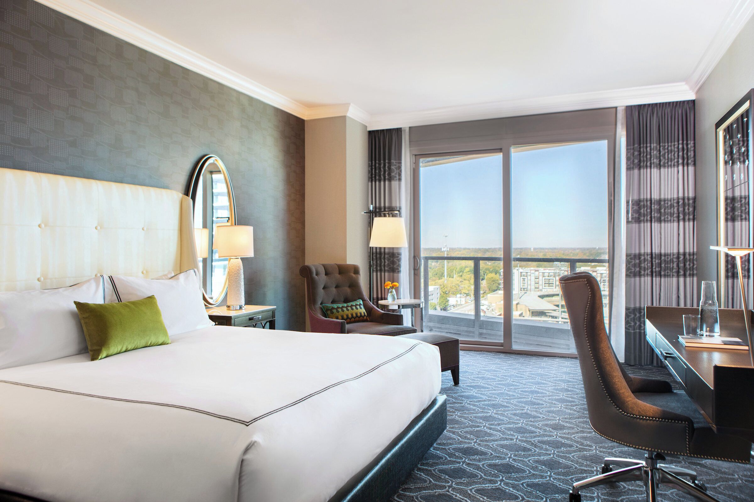 A hotel room with a large bed, a desk, a chair, and a view of the city through the windows.