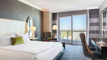 A hotel room with a large bed, a desk, a chair, and a view of the city through the windows.
