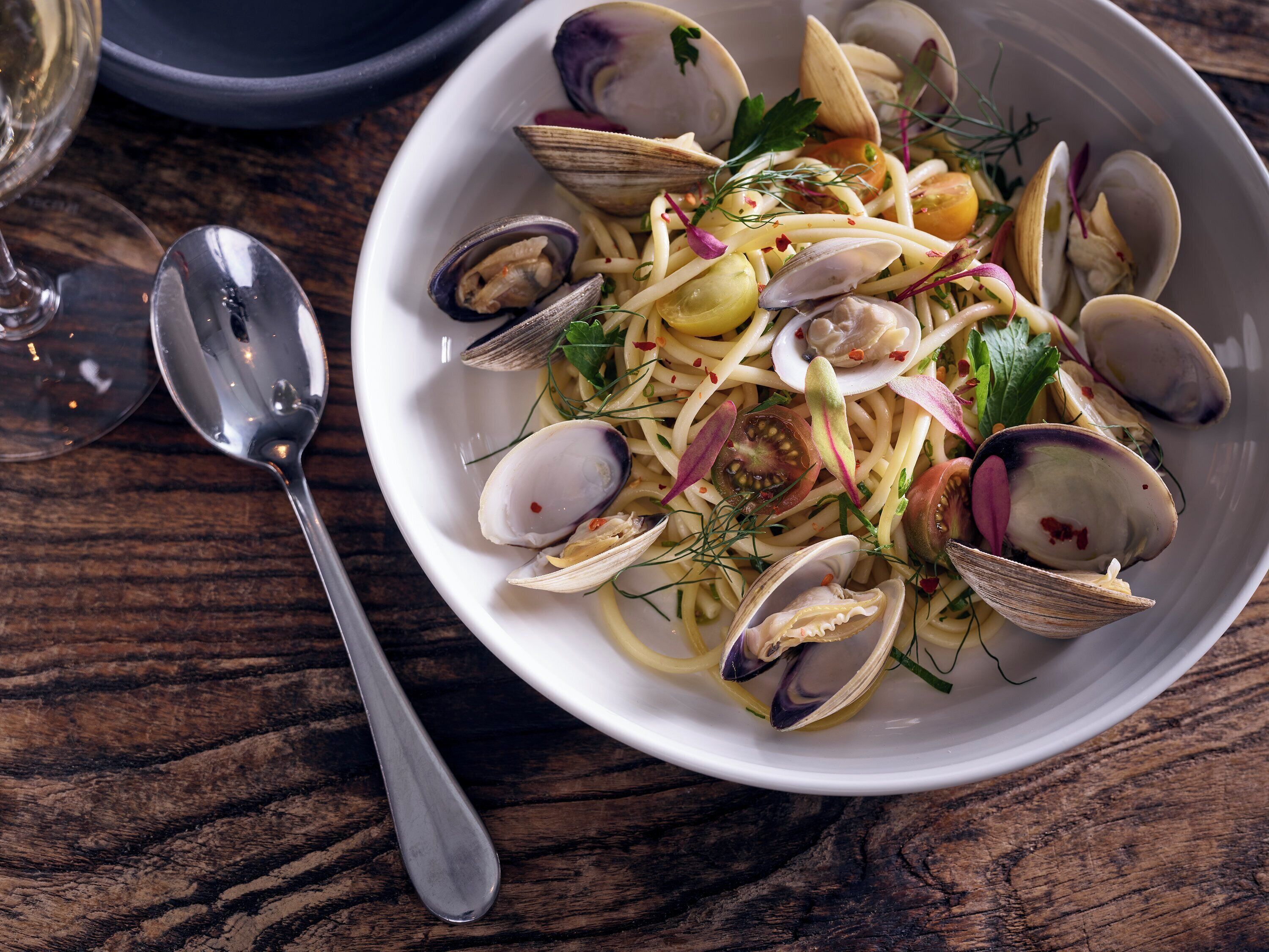A bowl of pasta with clams and vegetables on a wooden table.