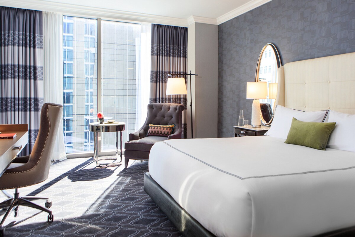A hotel room with a large bed, a desk, a chair, a small table, and a window with curtains.