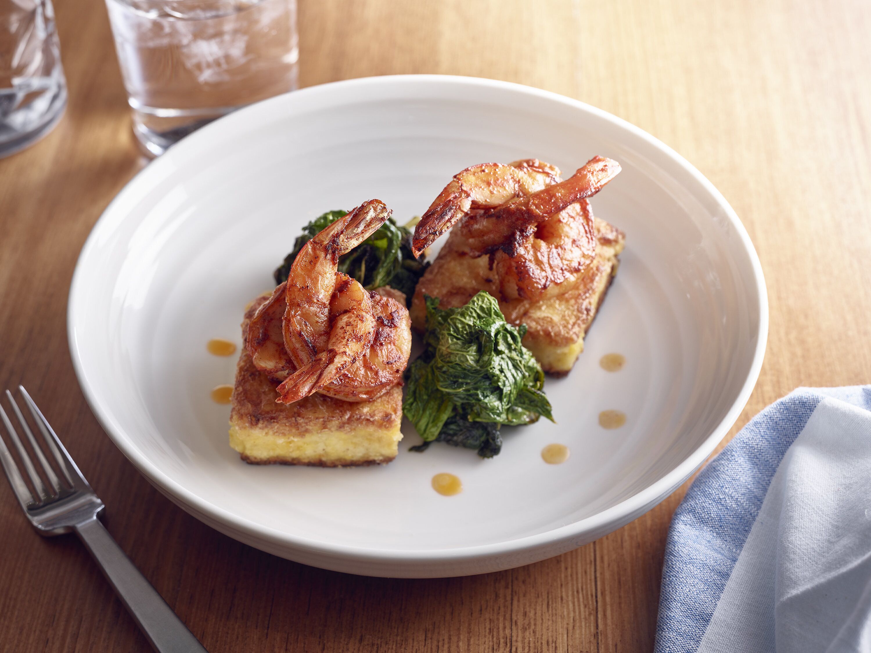 A white plate with a piece of food, a fork, and a glass of water on a wooden table.
