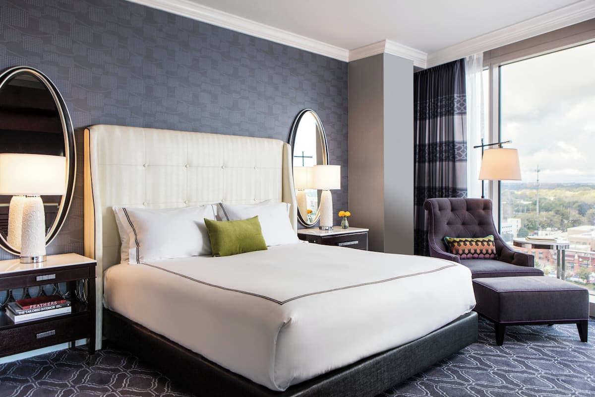Ein Hotelzimmer mit einem großen Bett, zwei Nachttischen mit Lampen, einem Sessel und Blick auf die Stadt.
