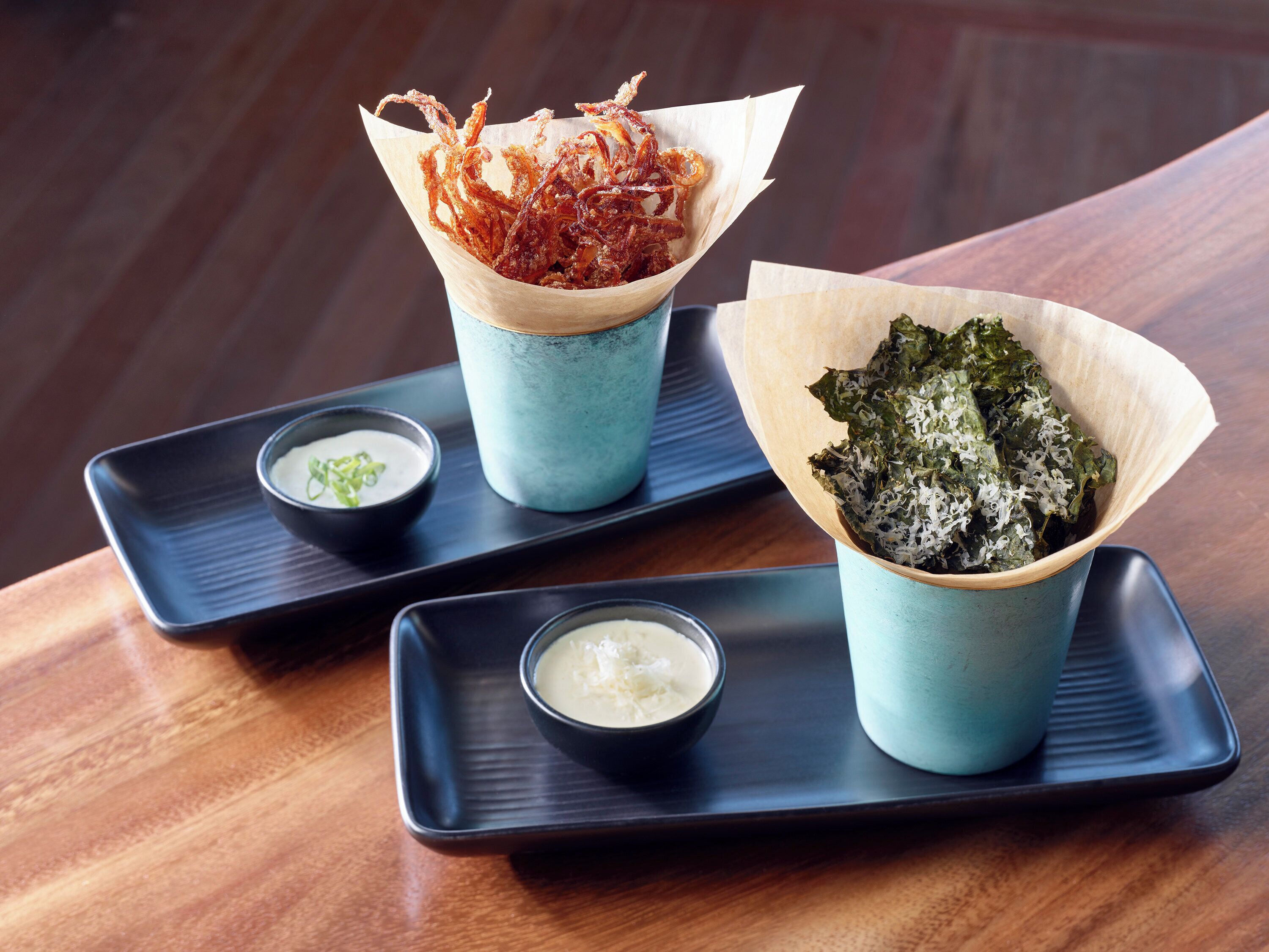 A tray with two bowls of food, one with fried items and the other with greens, and two small bowls of dipping sauces.