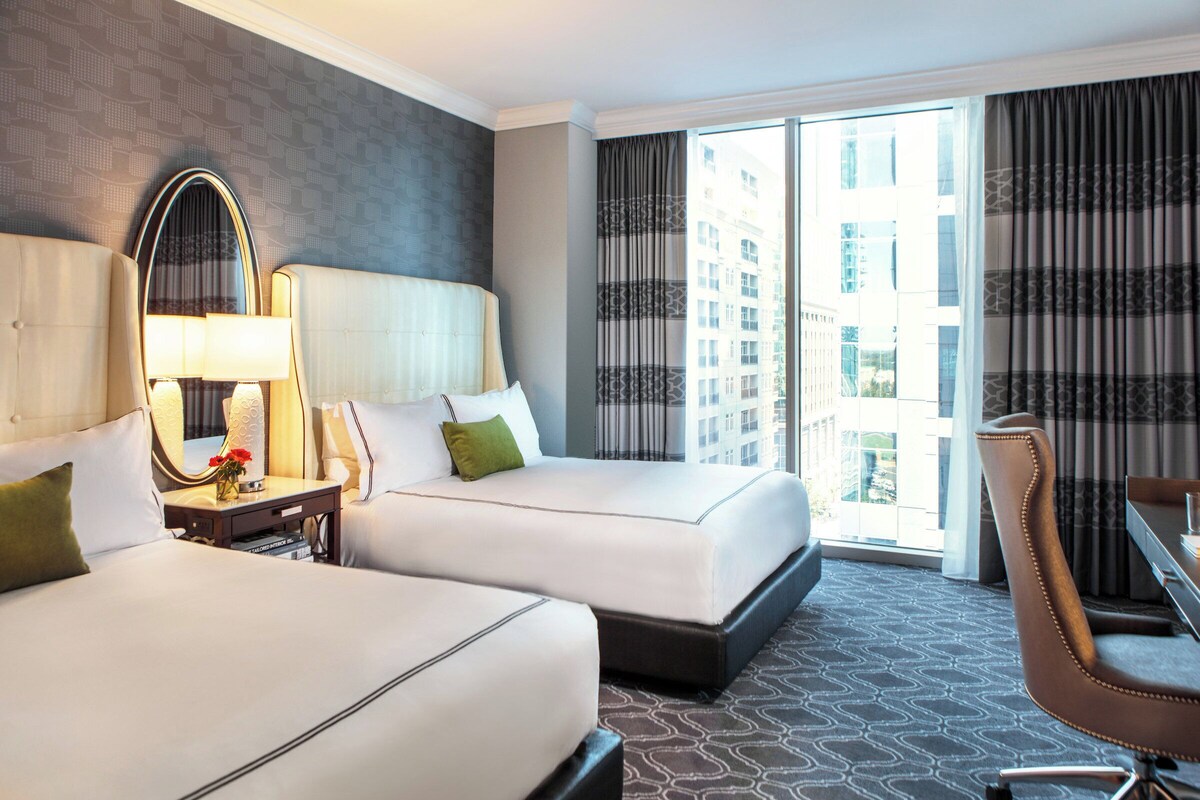 A hotel room with two beds, a desk, a chair, a large mirror, and a view of the city.