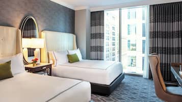 A hotel room with two beds, a desk, a chair, a large mirror, and a view of the city.