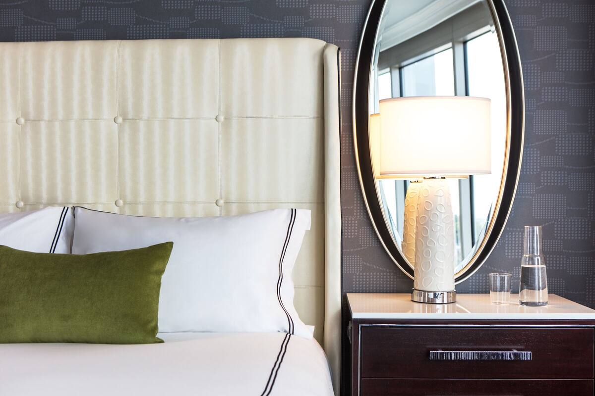 A hotel room with a tufted headboard, a bed with white and green pillows, a round mirror, a lamp, and a small table with a glass and a bottle.