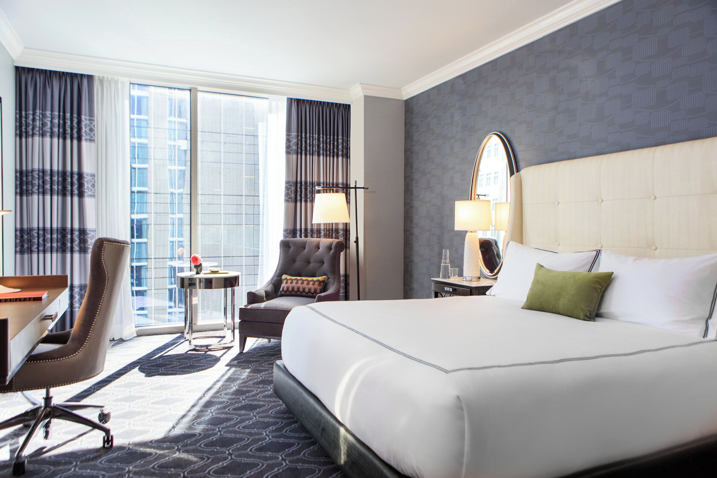 A hotel room with a large bed, a desk, a chair, a small table, and a window with curtains.