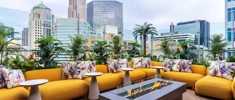 A rooftop lounge with yellow sofas, a glass-top coffee table, and a fire feature.