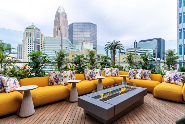 A rooftop lounge with yellow sofas, a glass-top coffee table, and a fire feature.