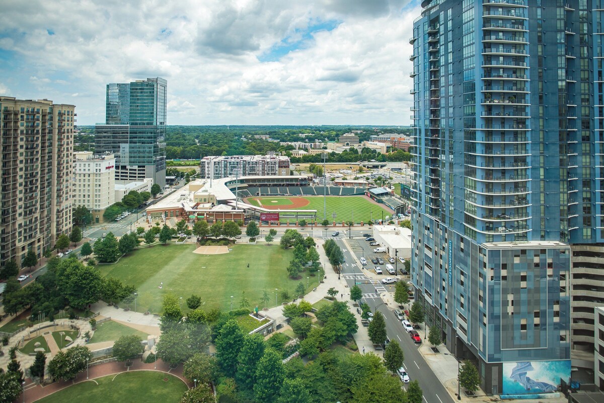 A high-rise building with a modern glass facade, surrounded by a well-maintained park with a baseball field, and adjacent to a parking structure.