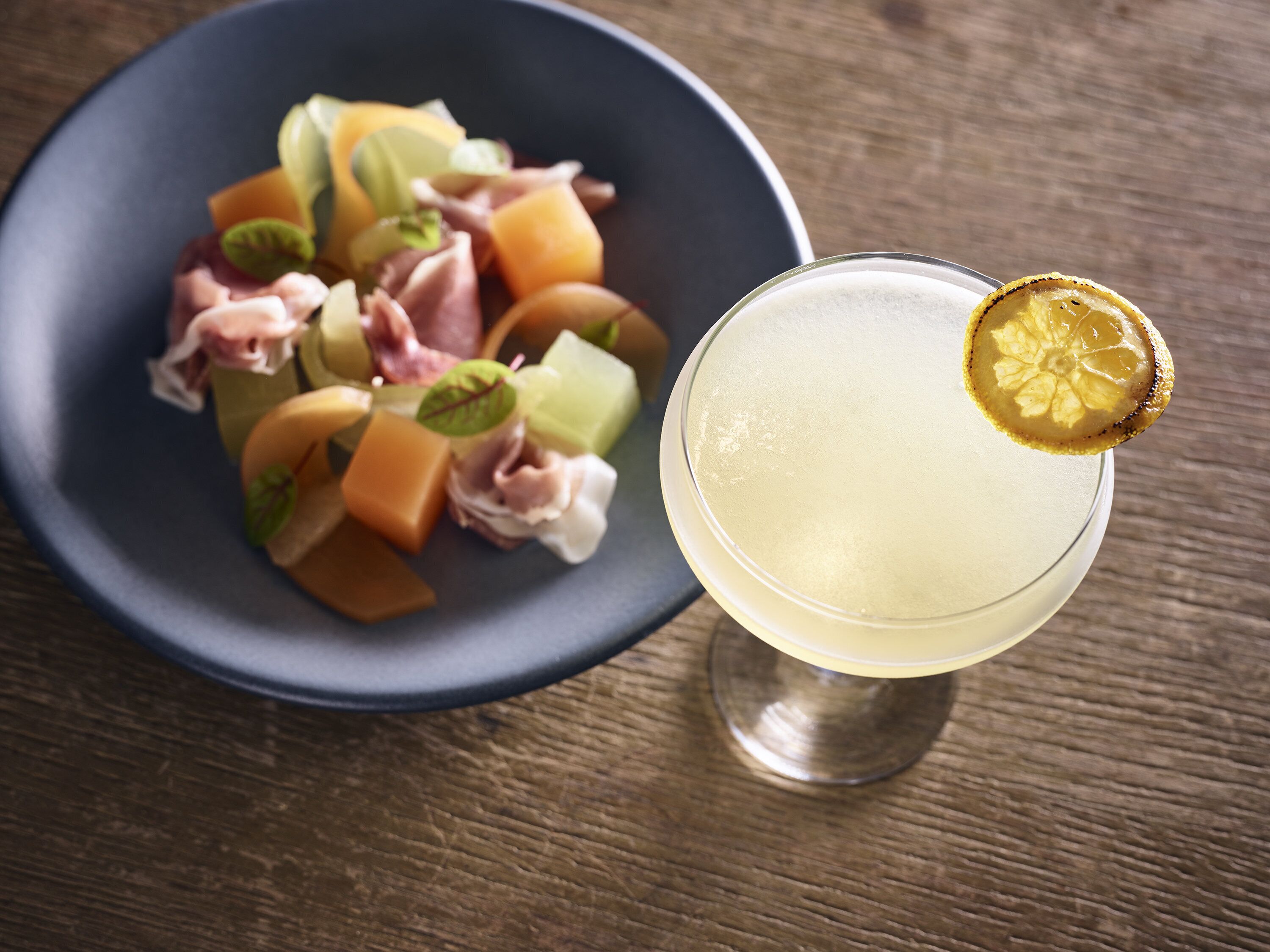 A dark blue bowl of fruit salad with a lemon garnish on a glass of clear liquid.
