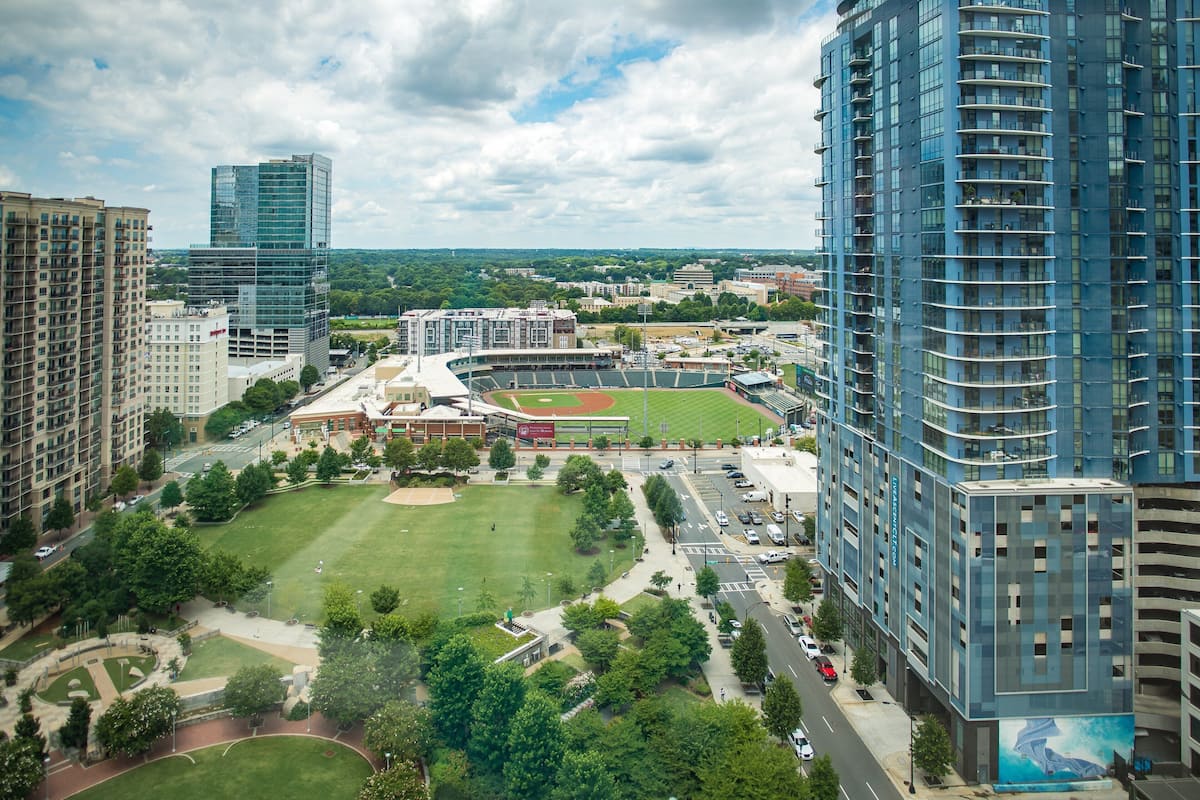 A high-rise building with a modern glass facade, surrounded by a well-maintained park with a baseball field, and adjacent to a parking structure.