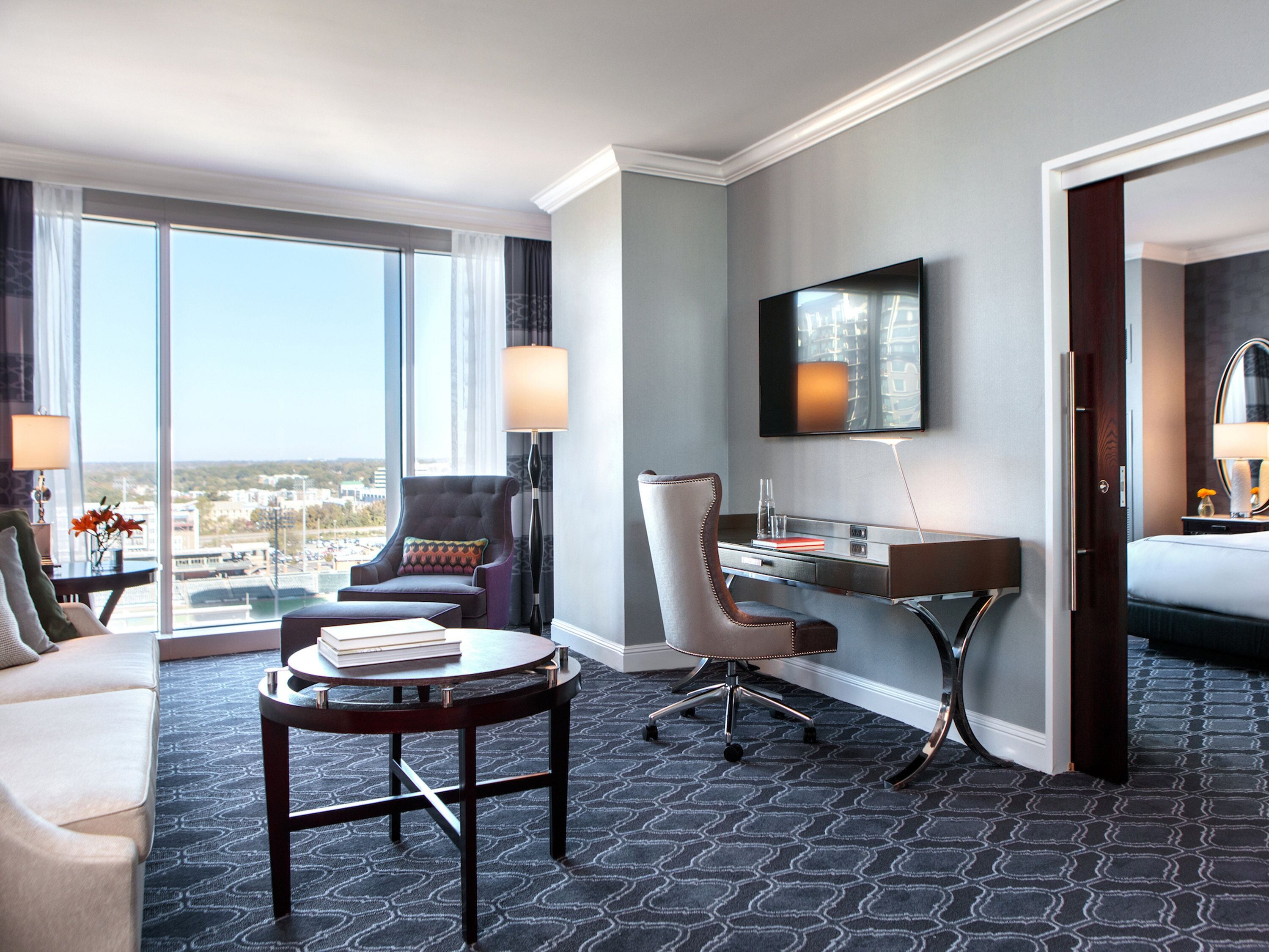 A hotel room with a large window, a desk, a chair, a sofa, and a television.
