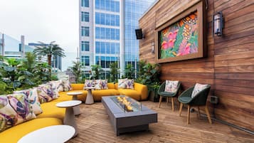 A rooftop lounge with yellow sofas, a glass-top table with a fire feature, and a wooden wall with a colorful mural.