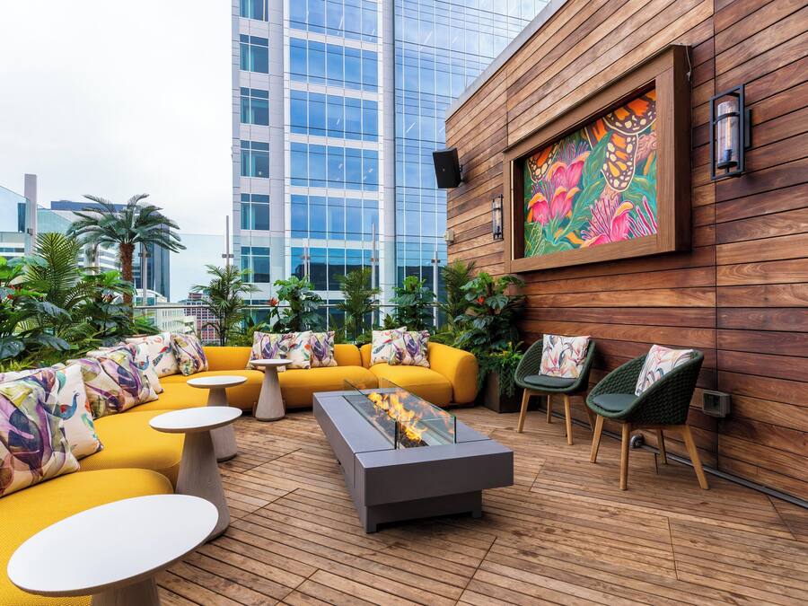 A rooftop lounge with yellow sofas, a glass-top table with a fire feature, and a wooden wall with a colorful mural.