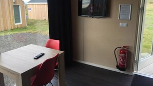Una habitación con una mesa de madera y sillas rojas, un televisor montado en la pared y un extintor cerca de la puerta.