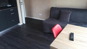 Un espacio de estar moderno con piso de madera oscura, un sofá negro y una silla roja, y una mesa de madera con un control remoto.