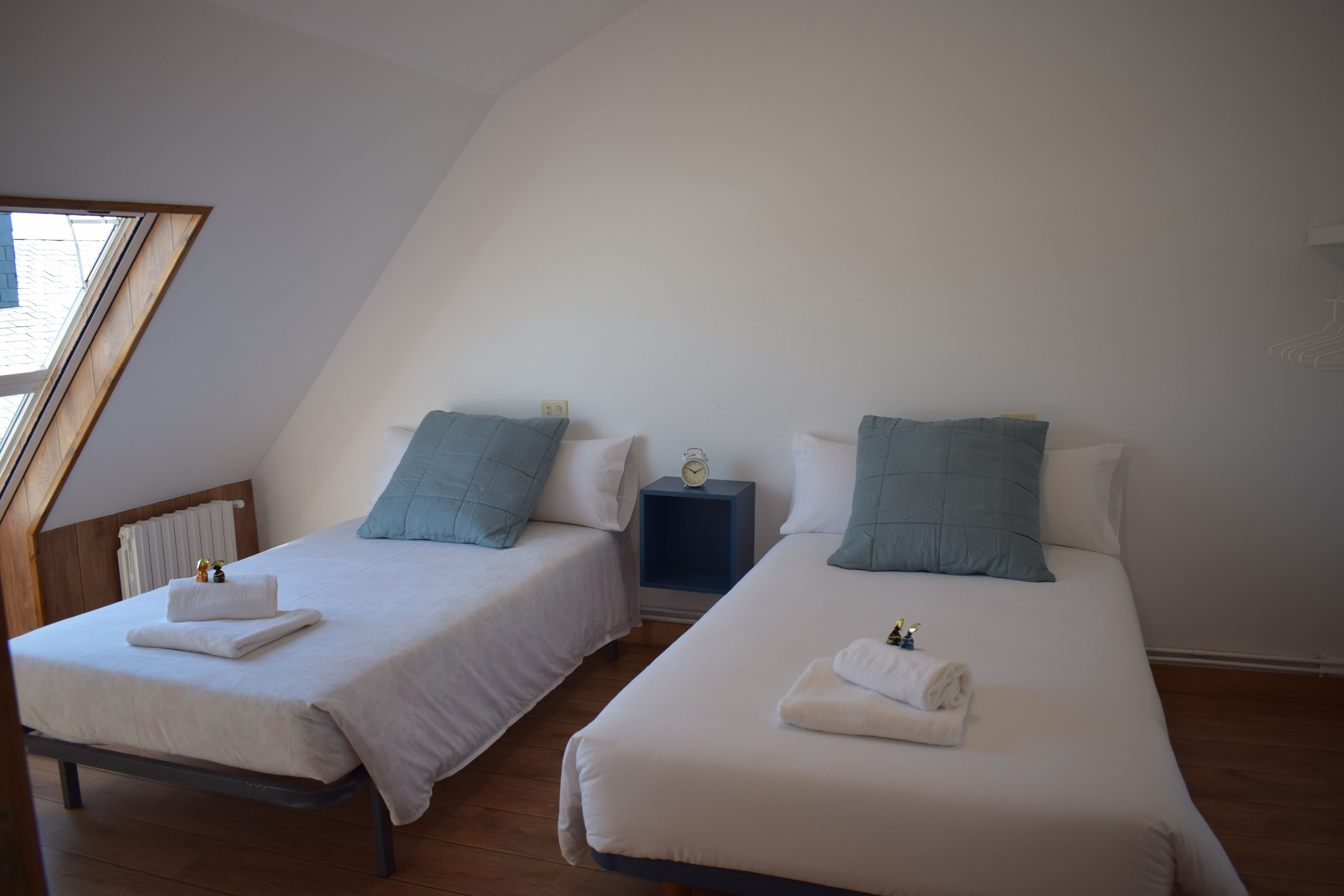 Dos camas individuales con sábanas blancas y almohadas azules, una mesita de noche con un reloj y una ventana con marco de madera.