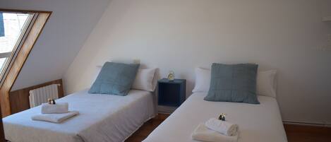 Dos camas individuales con sábanas blancas y almohadas azules, una mesita de noche con un reloj y una ventana con marco de madera.
