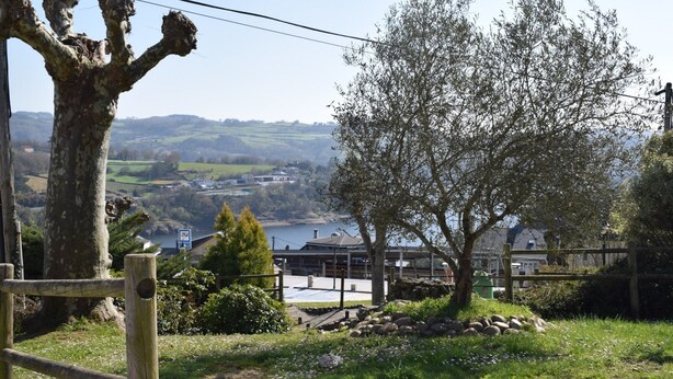 A view of a rural landscape with a large tree, a wooden fence, and a distant building by a body of water. - La Casa de Manuel (Portomarin)