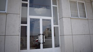 A building facade with a large glass window, a door with a number "12" above it, and a ventilation grille on the right side. - La Casa de Manuel (Portomarin)