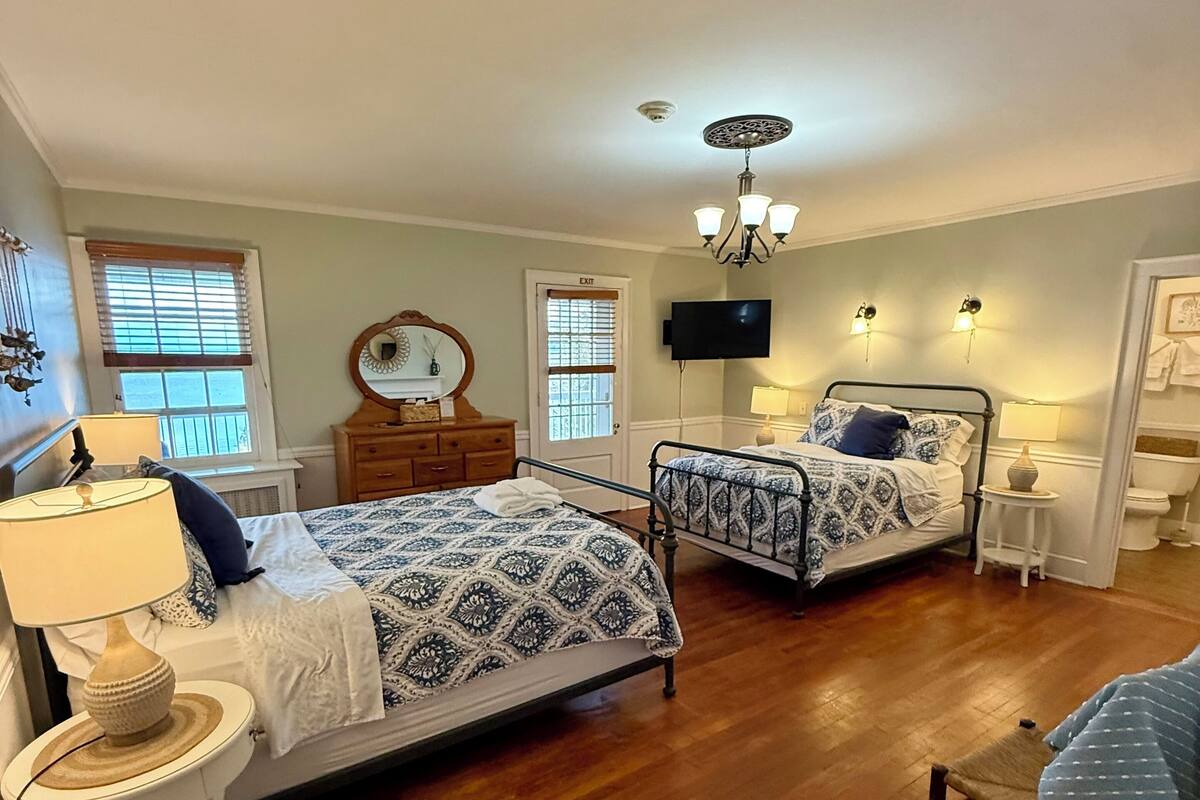Quarto com duas camas, cômoda, espelho, televisão e lustre.
