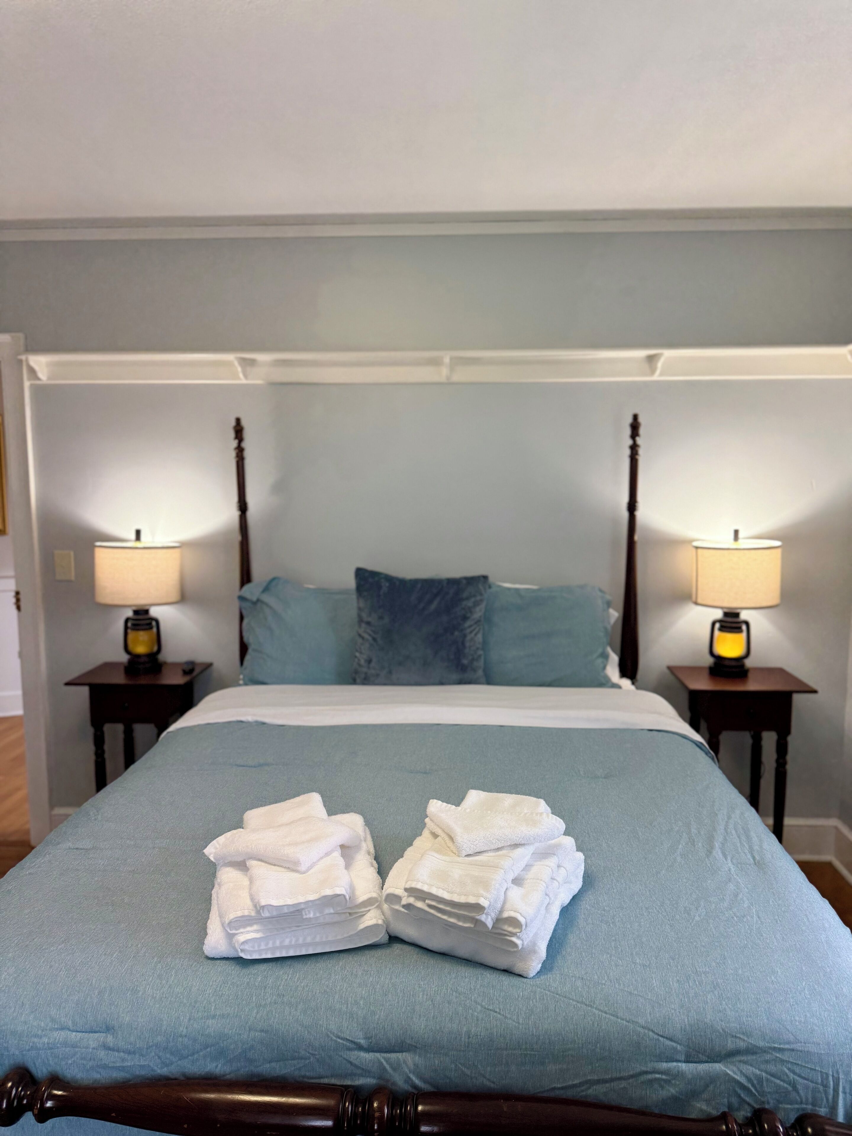Un lit bien fait, recouvert d’une couverture bleue, avec deux oreillers et une pile de serviettes blanches posée sur le pied de lit. On y trouve deux tables de chevet avec des lampes et une tête de lit en bois.