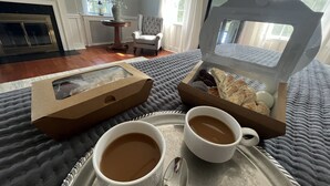 A cozy room with a bed, a tray with two cups of coffee, a box of pastries, and a vase with flowers.