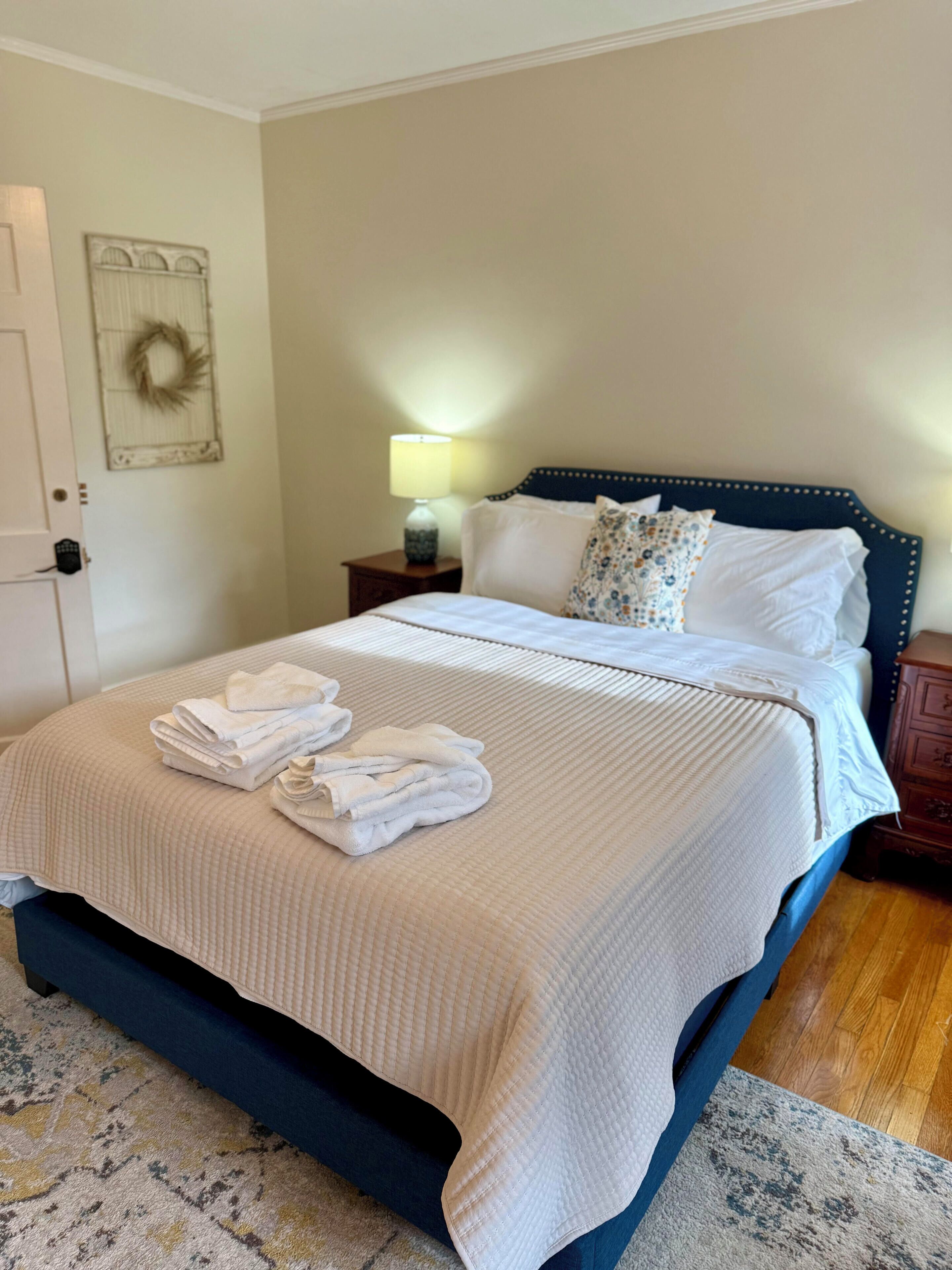A neatly made bed with folded towels, a bedside lamp, and a decorative wreath on the wall.
