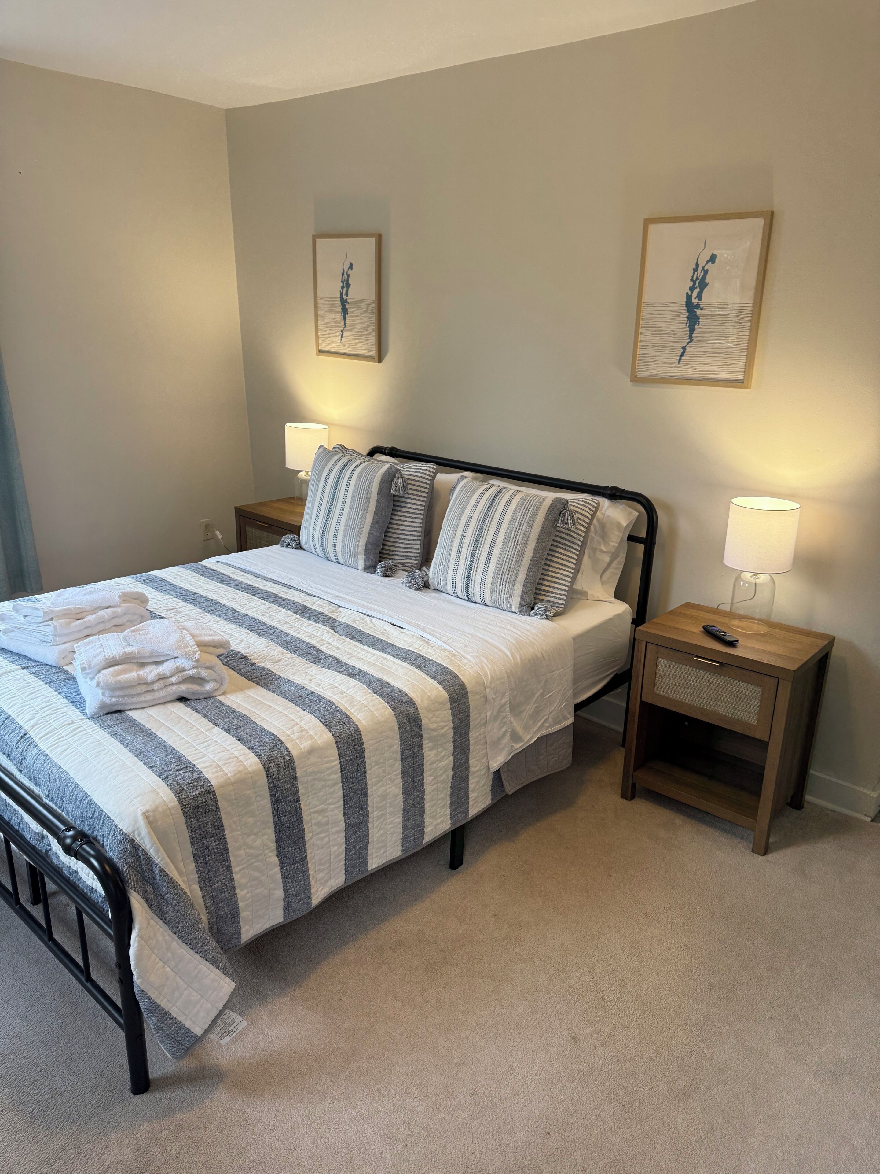 A neatly made bed with striped bedding and two pillows, a bedside table with a lamp, and two wall-mounted paintings.