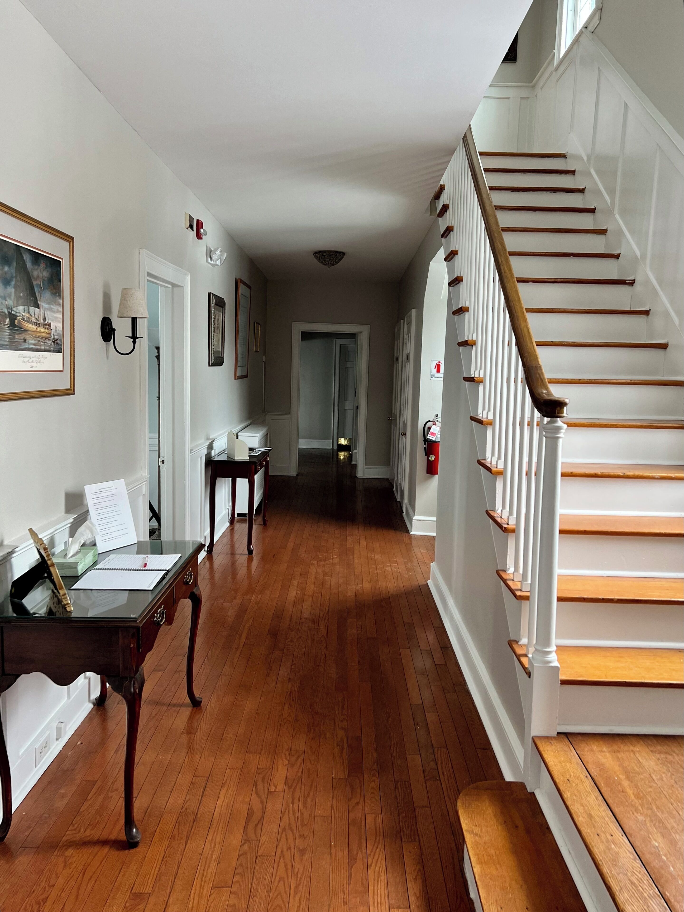 A hallway with wooden flooring, a staircase with white railings, and a desk with papers and a pen.