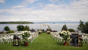 A wedding ceremony setup by a lake with chairs arranged in rows, a floral arch, and a chalkboard sign.