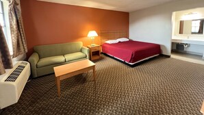 A hotel room with a bed, a green sofa, a small table, a nightstand with a lamp, and a bathroom with a mirror and sink. - Cotton Valley Motel (Clint)