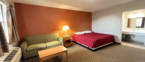 A hotel room with a bed, a green sofa, a small table, a nightstand with a lamp, and a bathroom with a mirror and sink.