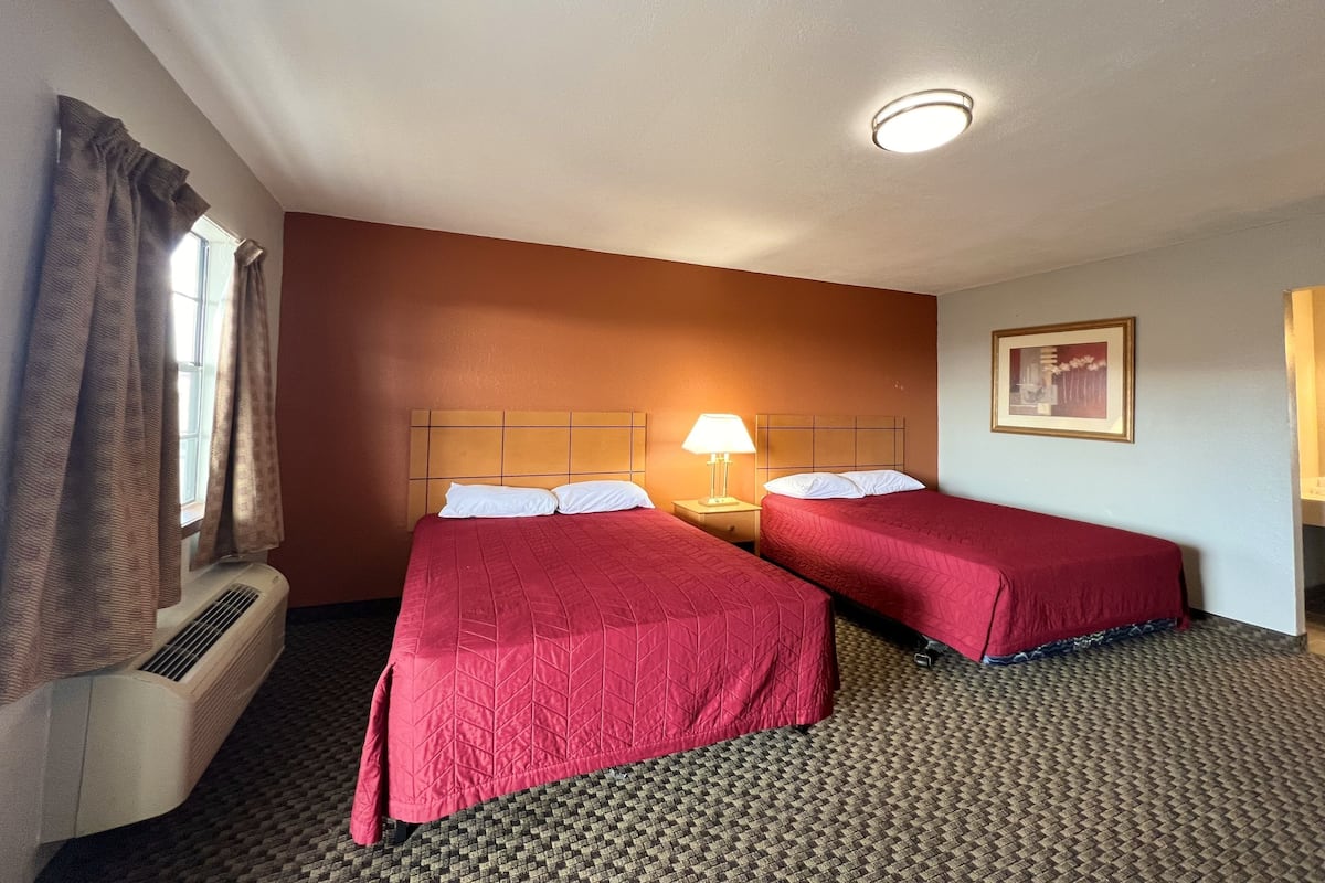 A hotel room with two beds, a heater, a window with curtains, a nightstand with a lamp, and a framed picture on the wall.