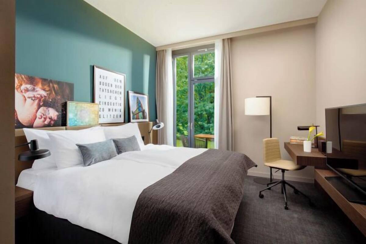 A hotel room with a bed, a desk, a chair, a TV, and artwork on the walls.