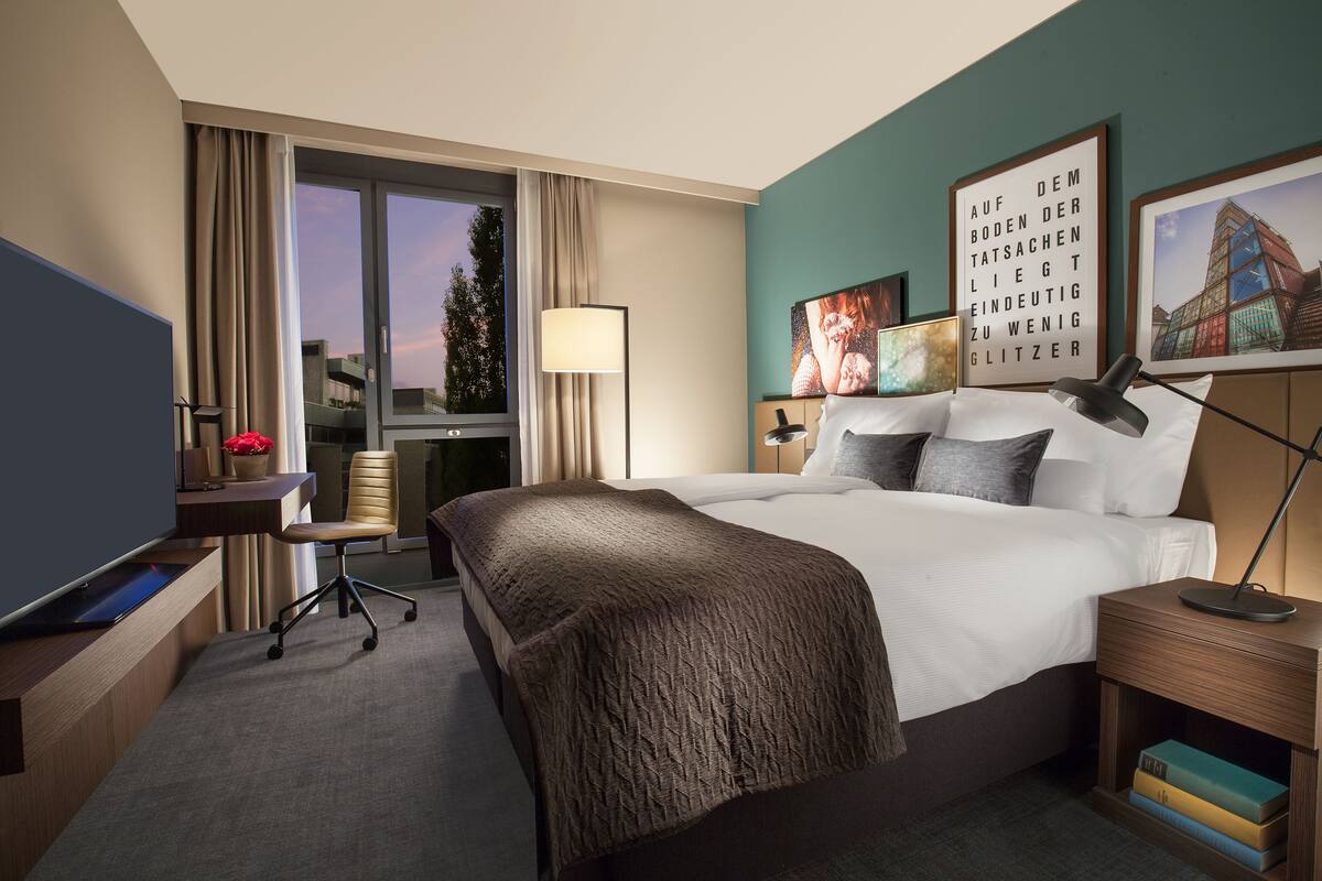 A hotel room with a bed, a desk with a laptop, a chair, a lamp, and a window with curtains.