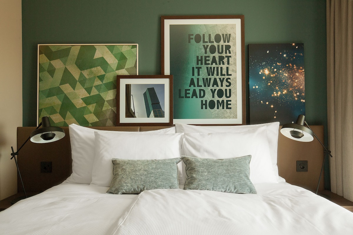 A neatly made bed with white linens and two pillows, flanked by two bedside tables with lamps, in a room with green walls and framed artwork.