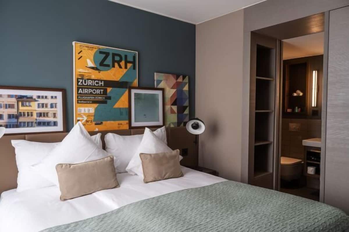 A hotel room with a bed, pillows, a wall-mounted poster, a bedside lamp, and a view into the bathroom.