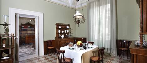 A dining room with a large table set for four, surrounded by wooden chairs. A chandelier hangs from the ceiling, and a wooden cabinet with porcelain tea sets is visible in the background.