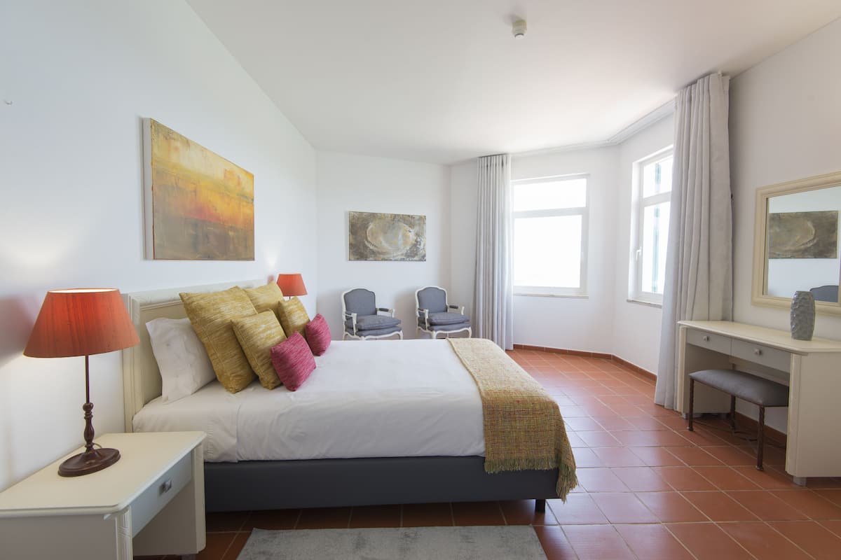 A bedroom with a bed, bedside table, lamp, two chairs, a desk, and a mirror.