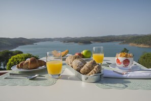 Una mesa de desayuno con cruasanes, fruta y vasos de jugo, con vista a un lago pintoresco.