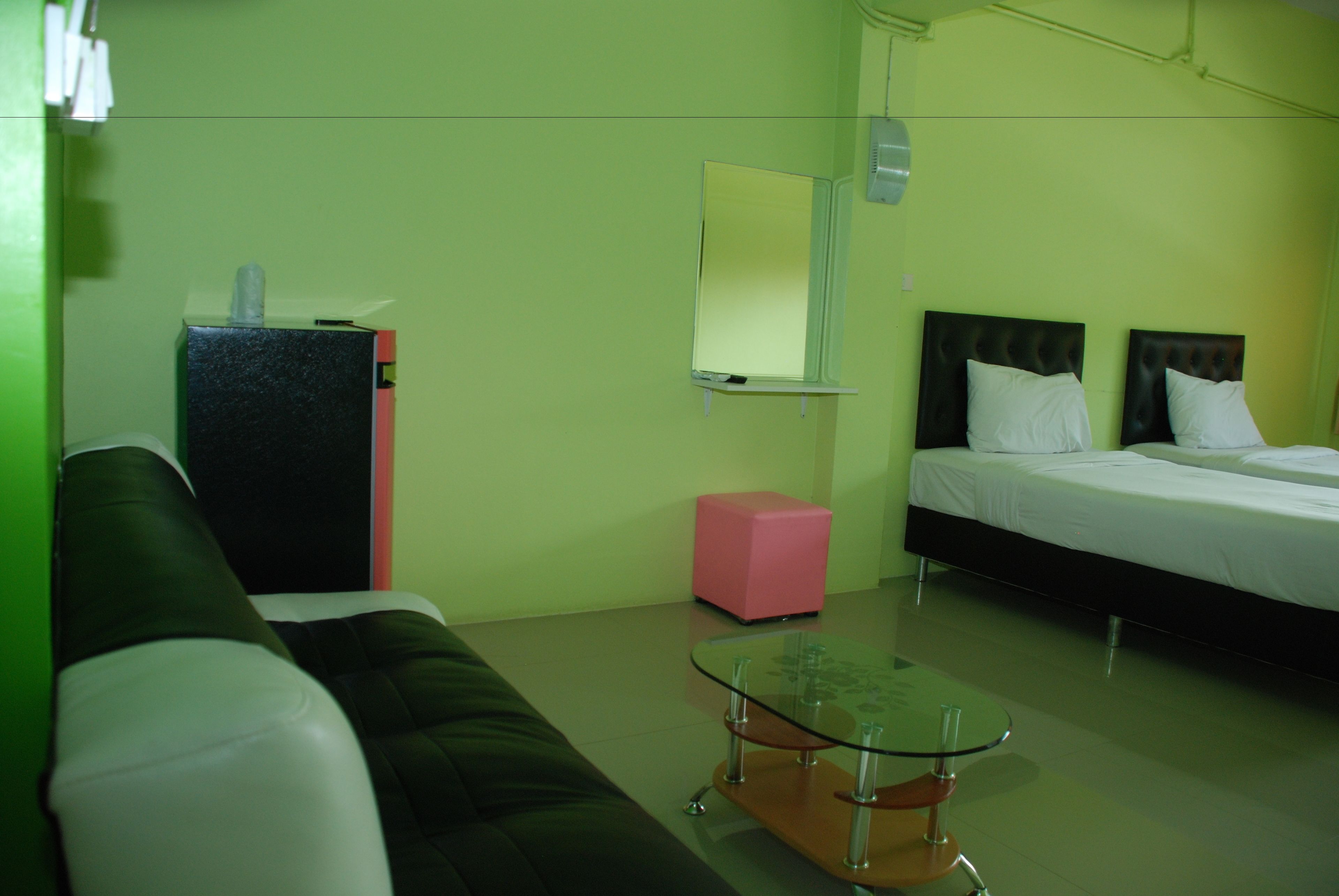 Habitación de hotel con una pared verde, una cama con ropa blanca, un cabecero negro, una mesa de centro de cristal, un reposapiés rosa y un sofá blanco y negro.