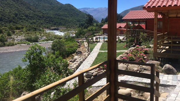 A wooden deck overlooking a river, with a view of mountains and a red-roofed building in the background. - Los Quenes River Lodge (Romeral)