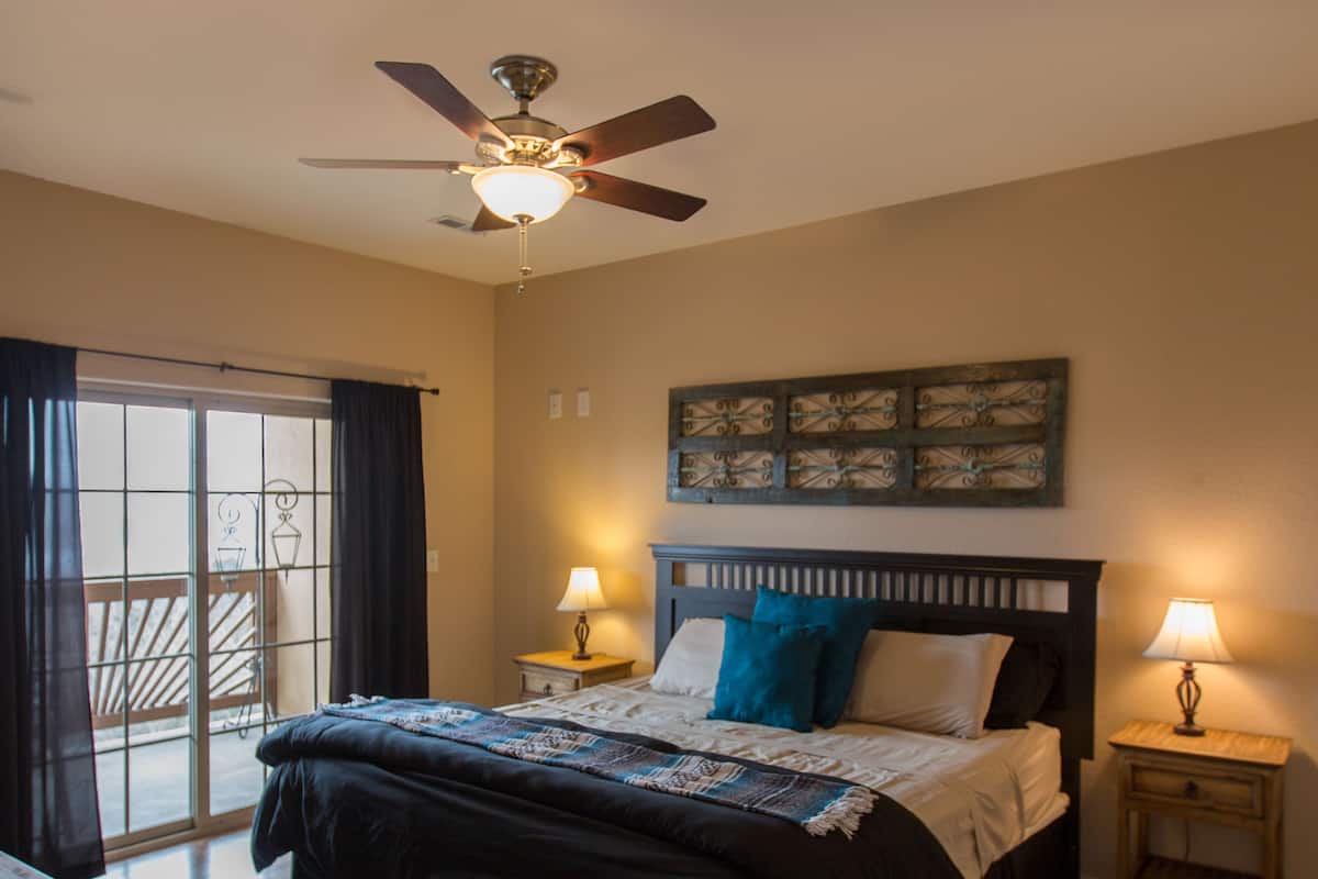 A bedroom with a bed, two bedside tables with lamps, a ceiling fan, a large window with curtains, and a wooden floor.