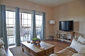 Ein Wohnzimmer mit Couch, Couchtisch, Fernseher auf einem Schrank und Blick durch eine Glastür auf einen Außenbereich mit Tisch und Stühlen.