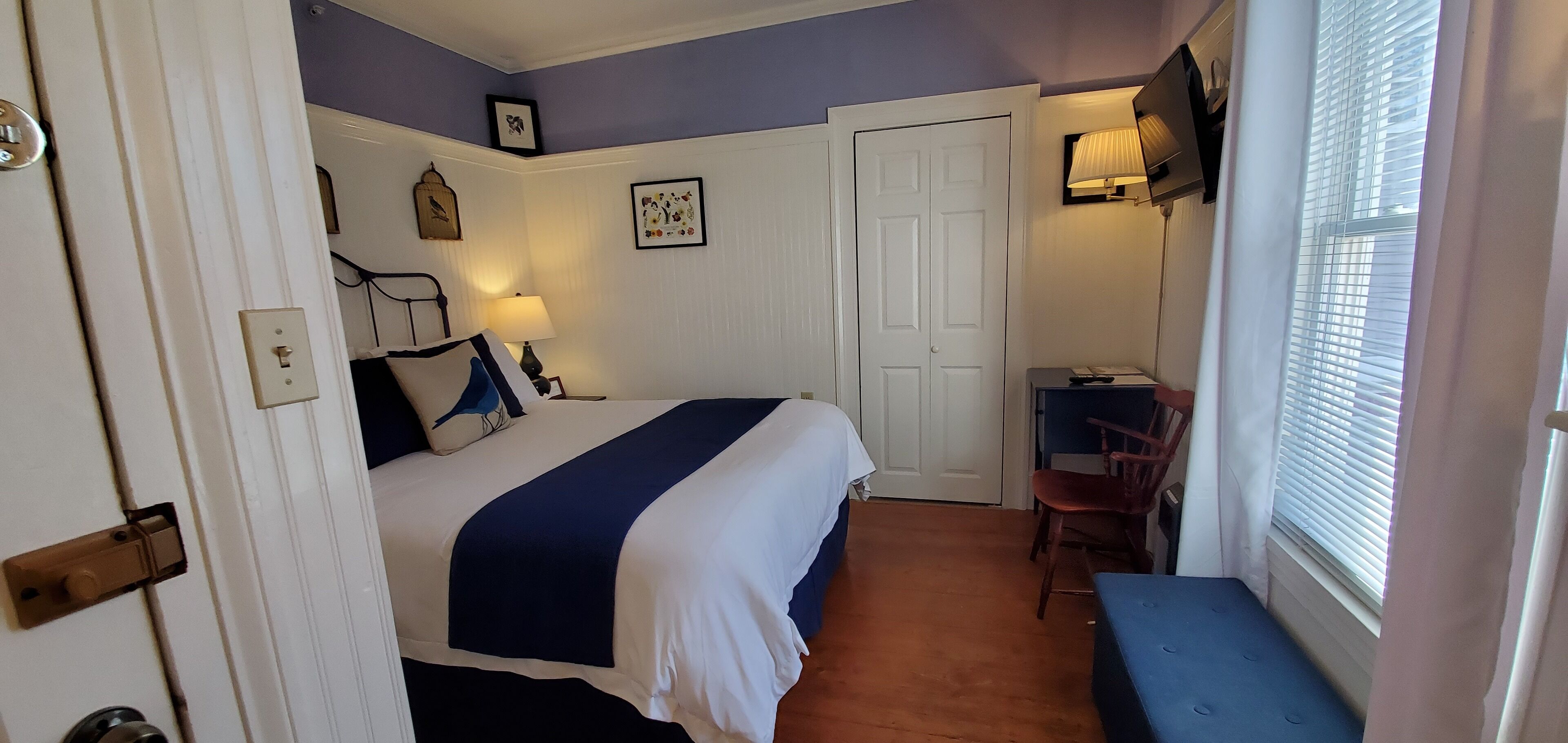 A bedroom with a bed, bedside table, lamp, chair, and a window with blinds.