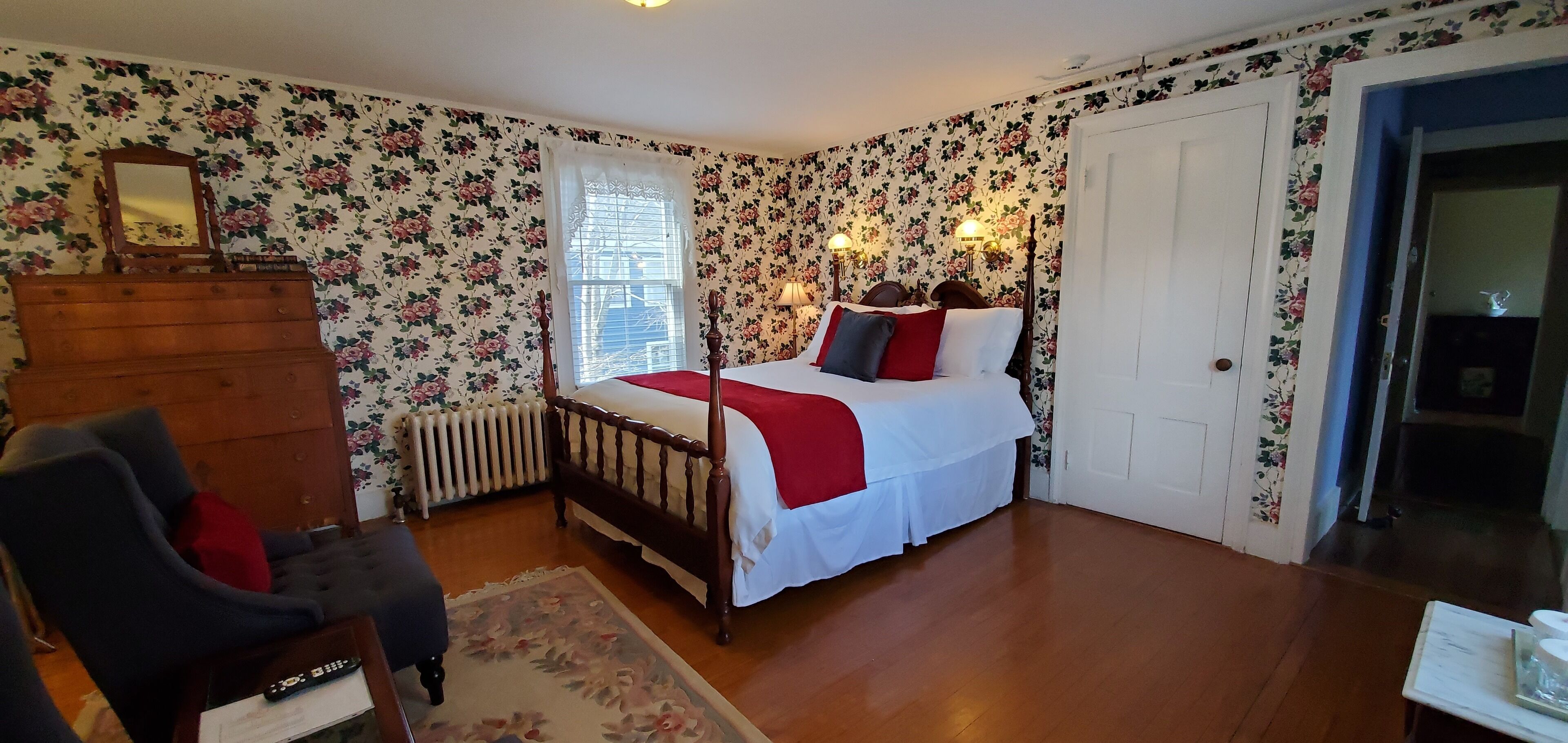 A bedroom with floral wallpaper, a wooden dresser, a bed with red and white bedding, and a nightstand.